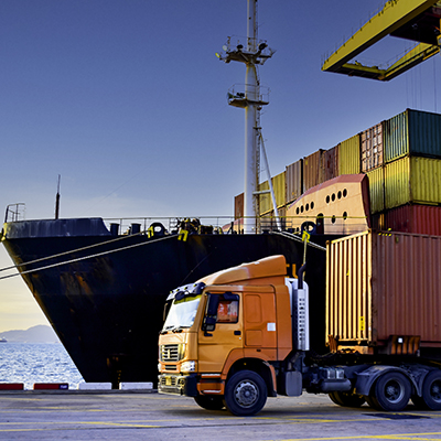 Truck carrying forty-foot container leaving port terminal with ship and quay crane on the background. Seaport operation activities, container shipping, and logistics.